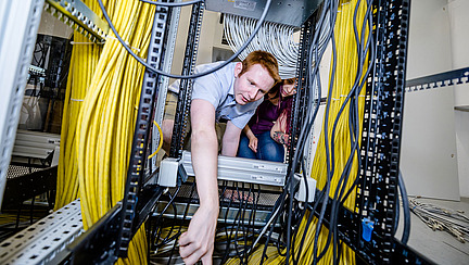 Der Betrachter befindet sich in einem Stahl-Regal mit vielen Kabeln. Eine Person lehnt sich von au&szlig;en hinein.