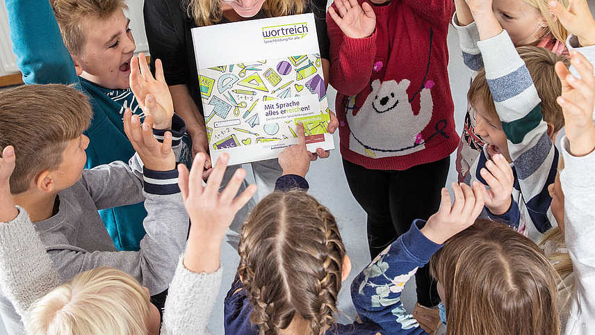 Mehrere Schüler und Schülerinnen stehen im Kreis und heben einen Arm. Eine Lehrerin hält eine wortreich Mappe hoch.