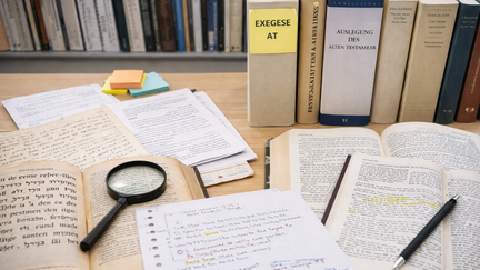 Ein Schreibtisch mit vielen aufgeschlagenen B&uuml;chern und Notizen. Mehrere dicke B&uuml;cher stehen auch aufrecht im Hintergrund, eines tr&auml;gt den Titel &bdquo;Exegese AT&ldquo;. Auf dem Tisch liegen offene Seiten mit markierten Textstellen. Daneben befinden sich handschriftliche Notizen auf losem Papier.  Eine Lupe liegt auf einem der B&uuml;cher, als w&uuml;rde jemand den Text genauer untersuchen. Ein Stift liegt bereit zum Schreiben. Insgesamt wirkt die Szene wie ein intensiver Arbeitsplatz zum Lesen, Lernen und Analysieren von Texten.