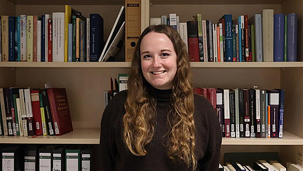 Eine junge Frau mit langen, welligen Haaren lächelt freundlich in die Kamera, umgeben von einem weißen Bücherregal voller Bücher und Ordnern. Sie trägt einen dunklen Pullover.