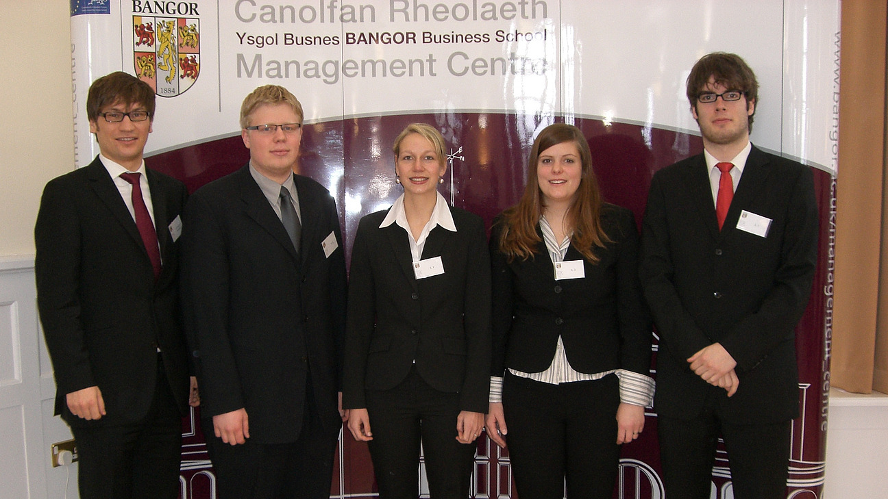 Drei M&auml;nner und zwei Frauen in Anz&uuml;gen vor einem Plakat der Bangor Business School stehend