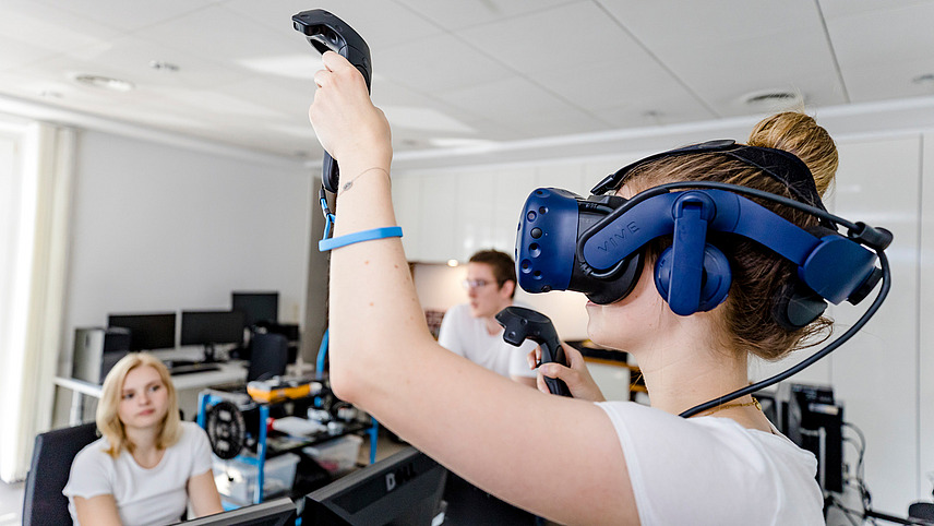 One student wears a VR headset and holds up a controller in her hand, another student sits at a computer in the background and observes her. Another student is standing at the technical equipment in the background.