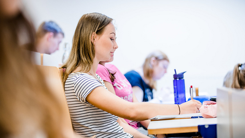 Side shot of a female student sitting on a bench in a lecture hall, taking notes on a paper pad. Other students are sitting around her.