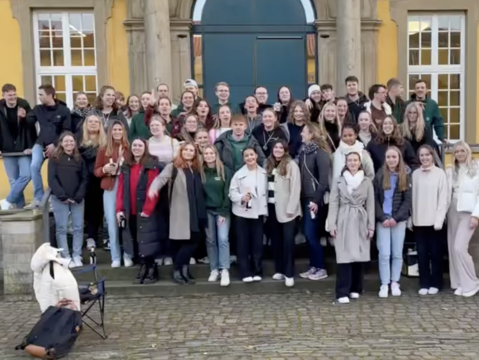 Eine gro&szlig;e Gruppe von Personen steht dicht nebeneinander auf einer Treppe vor einem Geb&auml;ude. Die Personen sind in mehreren Reihen angeordnet und schauen in Richtung Kamera.  Die Treppe f&uuml;hrt zu einer gro&szlig;en T&uuml;r, die von einer gelben Fassade mit Fenstern umgeben ist. Einige Personen tragen Jacken oder M&auml;ntel.  Im Vordergrund links ist ein Hund zu sehen. Die Gruppe f&uuml;llt den gesamten Bereich der Treppe aus.