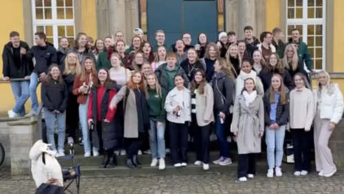 Eine gro&szlig;e Gruppe von Personen steht dicht nebeneinander auf einer Treppe vor einem Geb&auml;ude. Die Personen sind in mehreren Reihen angeordnet und schauen in Richtung Kamera.  Die Treppe f&uuml;hrt zu einer gro&szlig;en T&uuml;r, die von einer gelben Fassade mit Fenstern umgeben ist. Einige Personen tragen Jacken oder M&auml;ntel.  Im Vordergrund links ist ein Hund zu sehen. Die Gruppe f&uuml;llt den gesamten Bereich der Treppe aus.