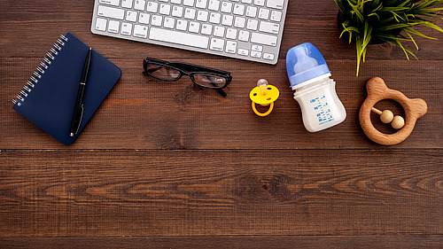 Work utensils lie on a desk next to a pacifier, a bottle and a baby toy.