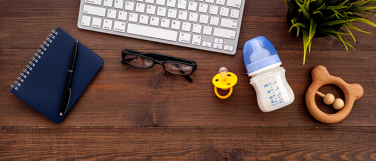 Work utensils lie on a desk next to a pacifier, a bottle and a baby toy.