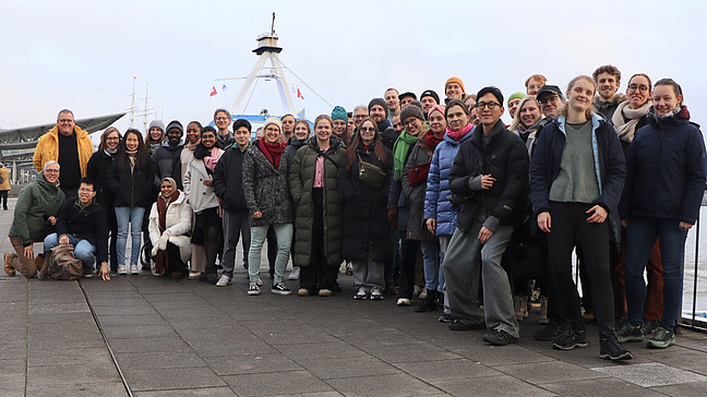 Eine Gruppe von 40 - 50 Personen l&auml;chelt in die Kamera, im Hintergrund sind Schiffe und Wasser zu sehen.