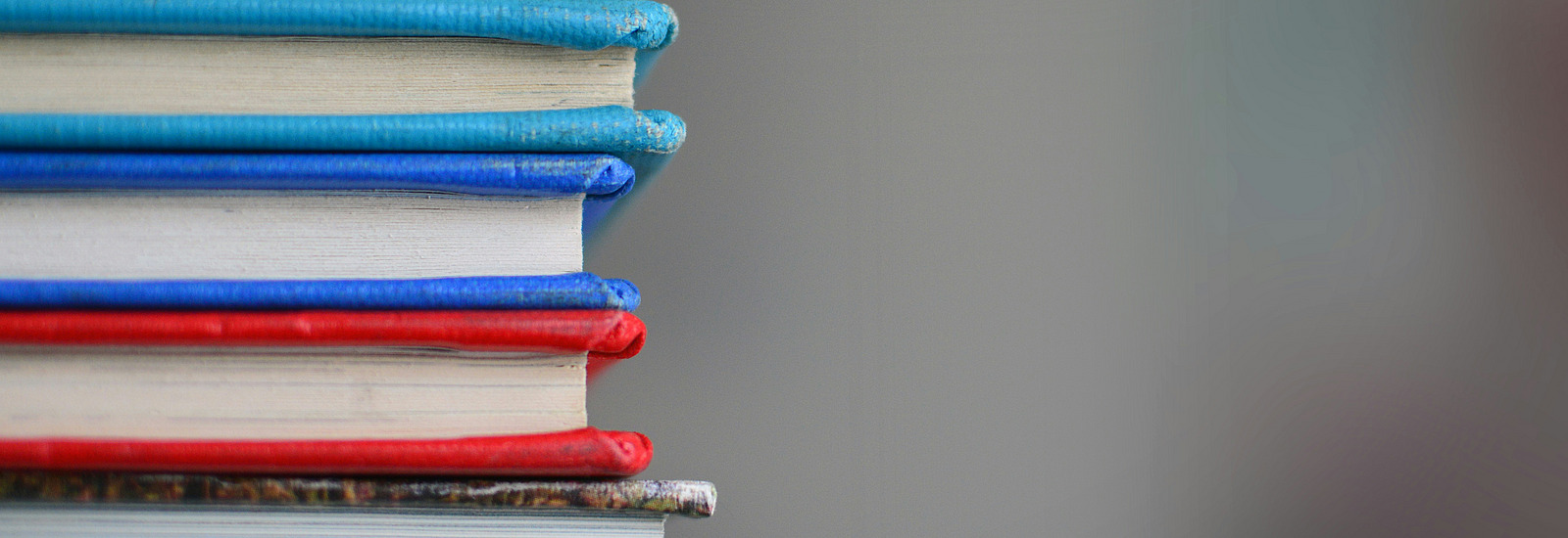 Sechs Bücher mit unterschiedlich farbigen Covern liegen aufeinandergestapelt. Der Hintergrund ist gräulich und unscharf.