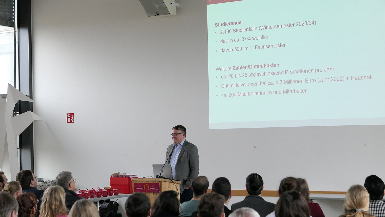 ../Download/Febr2024/P1000167.JPG The dean is standing behind a lectern. The image of the projector can be seen behind him.