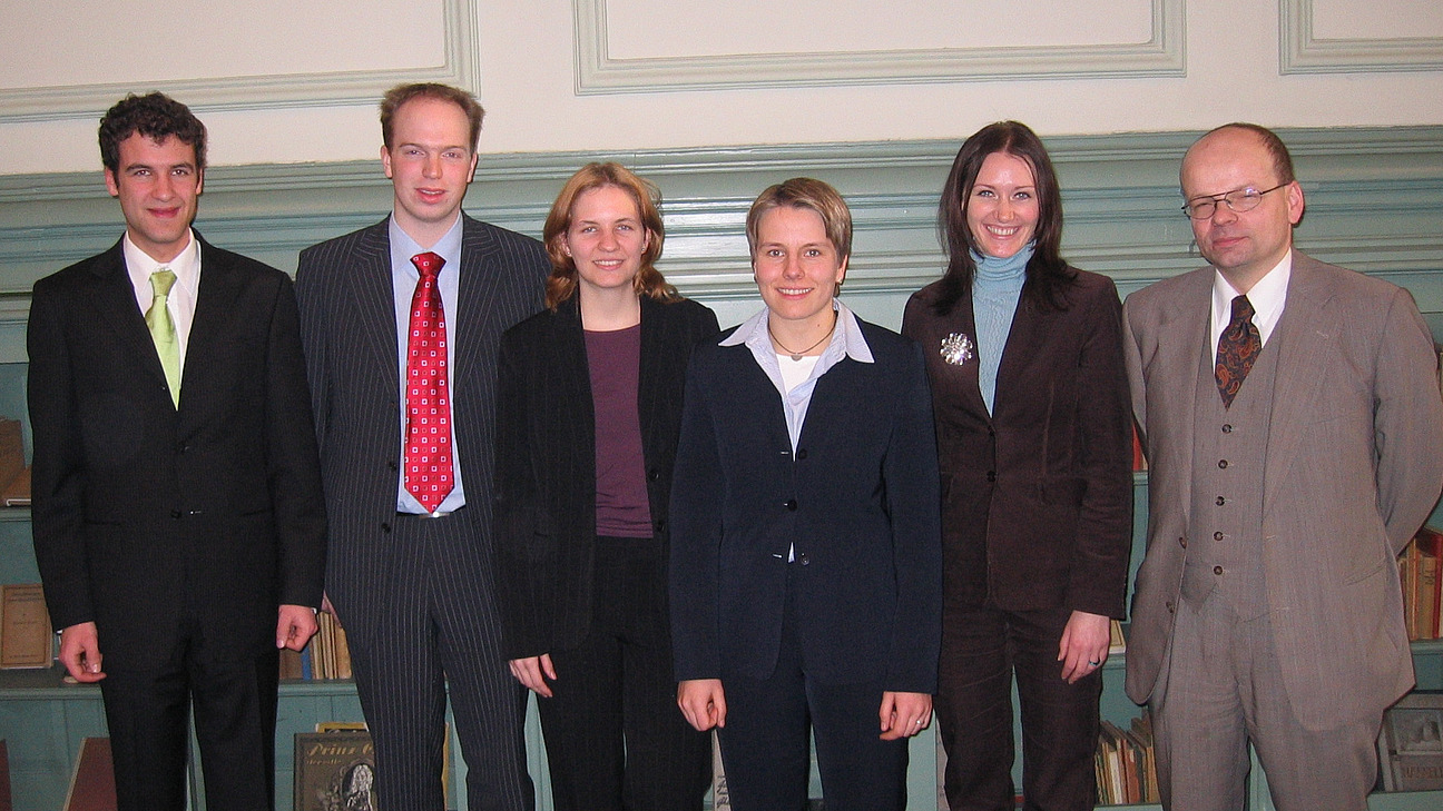 Drei M&auml;nner und drei Frauen in Anz&uuml;gen vor einer gr&uuml;nen holzvert&auml;felten Wand stehend 