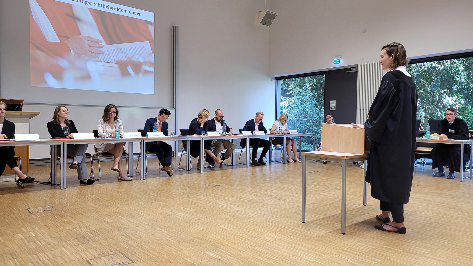 Frau in schwarzer Robe hinter Rednerpult vor einer Tischreihe mit sitzenden "Richtern" stehend