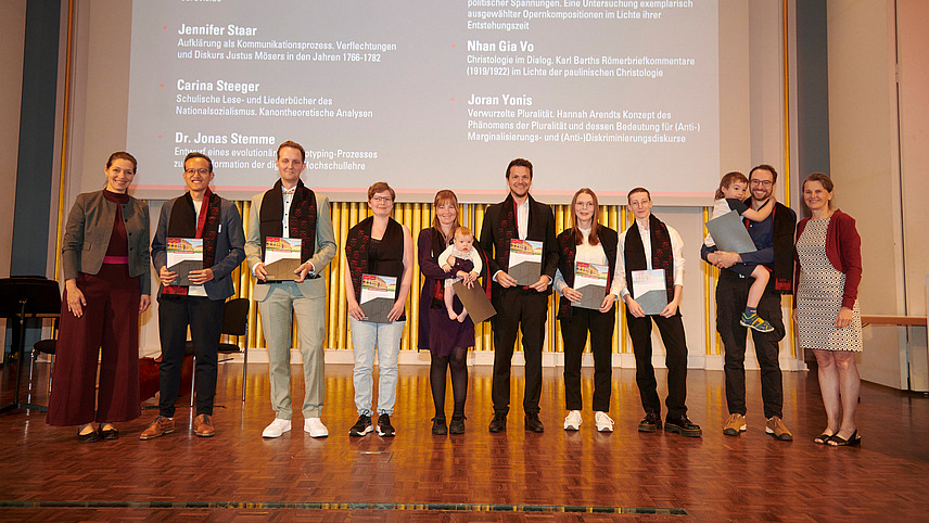 Several people stand on a stage, holding certificates in their hands and smiling into the camera