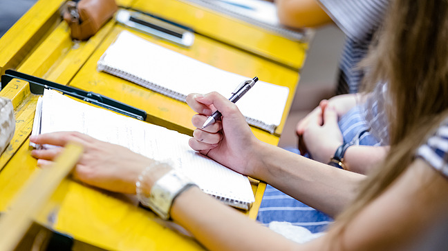Studierende sitzen in einem H&ouml;rsaal. Vor ihnen liegen Bl&ouml;cke.