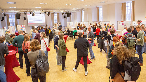 Viele Personen in einem großen Raum, an den Wänden stehen Stellwände