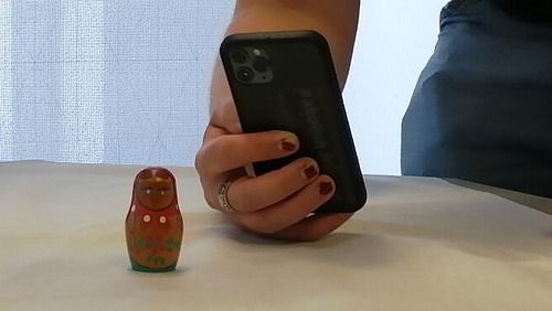 Das Bild zeigt eine Hand, die ein schwarzes Smartphone hält. Vor der Hand und dem Smartphone steht eine kleine, traditionelle russische Matroschka-Puppe. Die Matroschka ist bunt bemalt und hat rote Backen. Der Hintergrund ist ein weißer Tisch oder eine weiße Oberfläche. Die Person, die die Hand hält, trägt einen dunklen Rock und hat rot lackierte Fingernägel und einen Ring am Finger. 