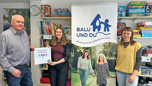 Drei Personen stehen vor eine Regal mit Spielen und Büchern, zwischen ihnen steht ein Banner von Balu und Du. Zwei Personen halten eine Urkunde mit der Fördersumme in die Kamera.