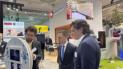 Three men stand next to each other and look at a screen.