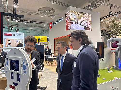 Three men stand next to each other and look at a screen.