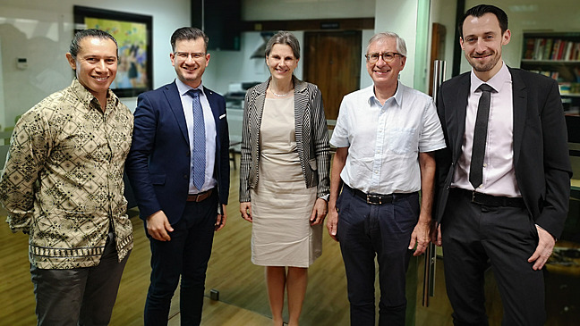 Das Bild zeigt eine Gruppe von f&uuml;nf Personen, die nebeneinander stehen. Im Hintergrund sieht man einen grossen Raum/Konferenzraum. Die Stimmung scheint aufgelockert und freundlich zu sein.
