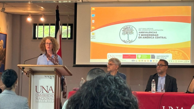 The image shows a panel discussion or presentation taking place. Several people are seated at a table behind a podium. A woman is speaking at the podium. The backdrop displays a presentation slide with text in Spanish.