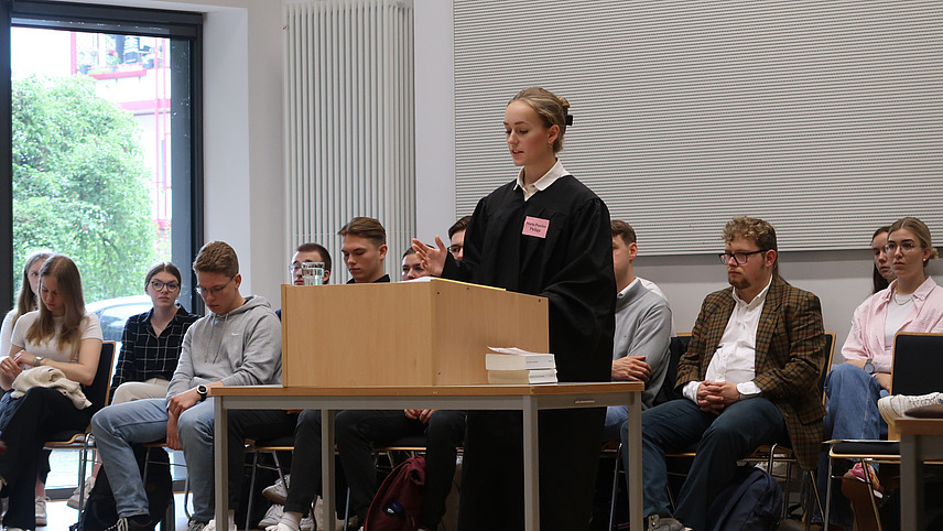 Junge Frau in schwarzer Robe hinter einem Rednerpult stehend, im Hintergrund sitzendes Publikum