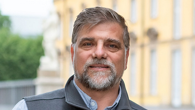 Nahaufnahme von Herrn Prof. Dr. Rass. Ein Mann mit grau meliertem Bart und freundlichem Blick. Er trägt eine Weste über einem karierten Hemd