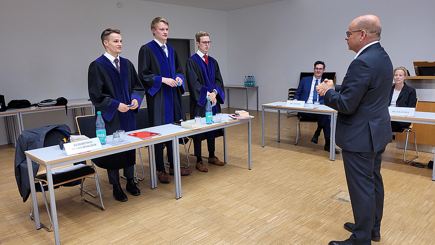 Professor D&ouml;rr in blauem Anzug spricht mit drei M&auml;nnern in schwarz-blauen Roben, die hinter einer Tischreihe stehen
