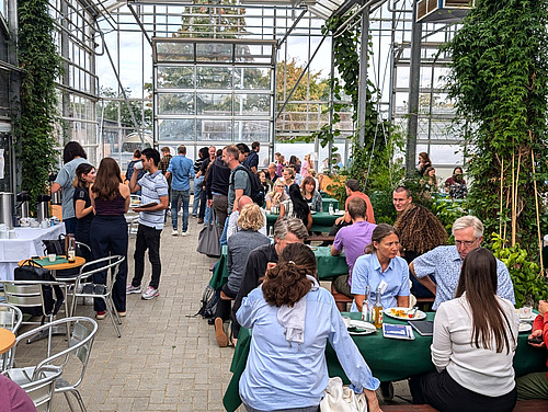 People are sitting and standing in a large greenhouse and talking to each other.