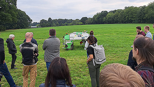 Auf einer grünen Wiese fährt ein autonomer Traktor. Im Vordergrund stehen 8 Personen und beobachten den Traktor.