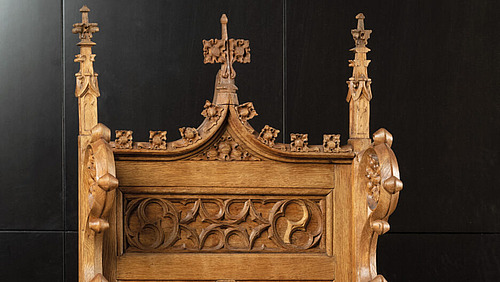 Ein h&ouml;lzernes Objekt mit aufwendigen Schnitzereien. Es ist hellbraun und zeigt oben mehrere spitze, turmartige Aufs&auml;tze.  Die Oberfl&auml;che ist mit Ornamenten und Mustern verziert. In der Mitte befindet sich ein quer verlaufendes Feld mit durchbrochenen Formen.  Der Hintergrund ist dunkel und hebt das Holzobjekt deutlich hervor.