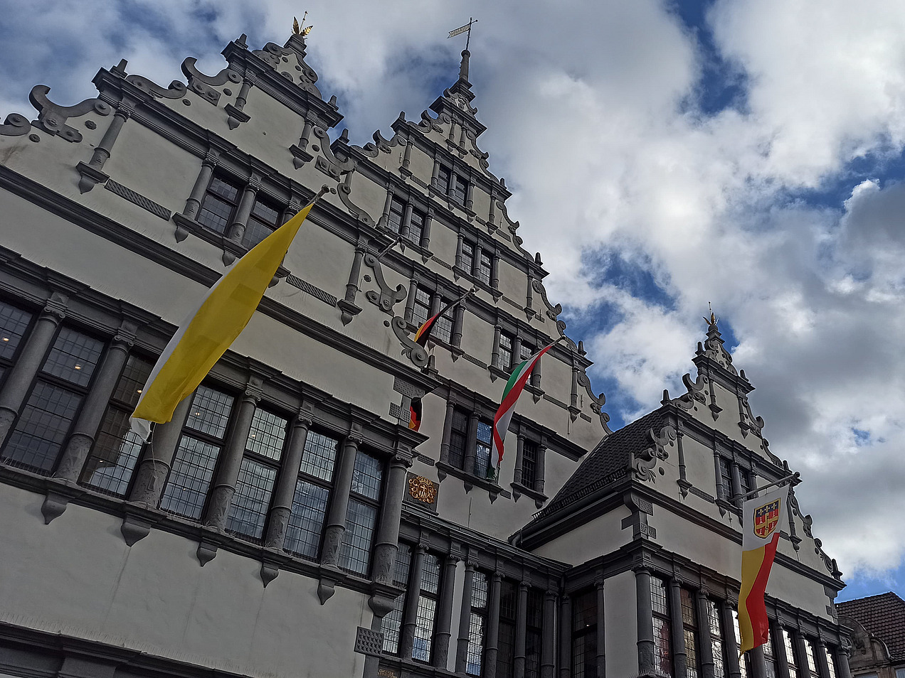 Das Bild zeigt ein weißes Haus mit drei Giebel. Die Fenster sind grau mit Stein umrandet und an ihnen hängen bunte Fahnen.