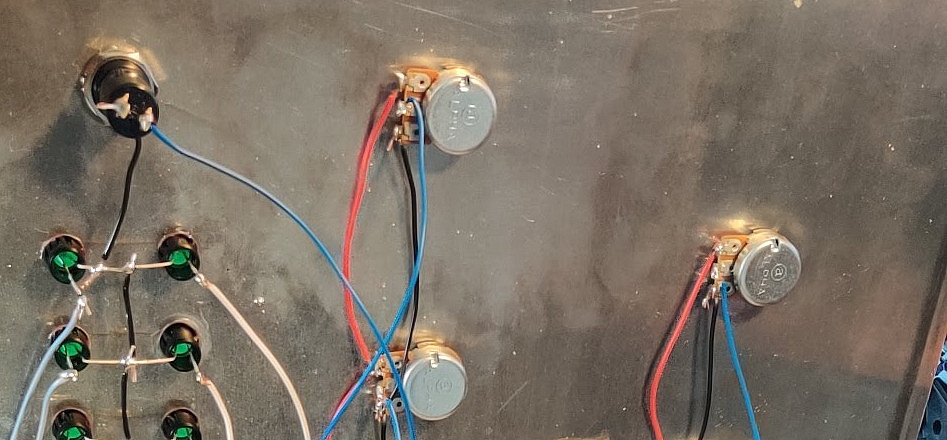 Das Bild zeigt das Innere eines elektronischen Geräts oder eines DIY-Projekts. Mehrere Potentiometer und Taster sind auf einer Metallplatte montiert, die mit verschiedenen farbigen Kabeln (blau, rot, schwarz und weiß) verbunden sind. Die Kabel verlaufen von den Bedienelementen zu einer Platine oder einem Breadboard, das am Boden eines Holzgehäuses befestigt ist.