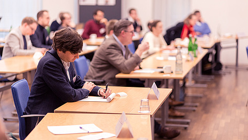 Mehrere Personen sitzen an Tischen in einem Raum und arbeiten mit Unterlagen. Im Vordergrund sitzt eine Person und schreibt auf ein Blatt Papier.  Auf den Tischen stehen Wasserflaschen, Gl&auml;ser und Namensschilder. Die Personen sitzen in Reihen und sind in Richtung einer gemeinsamen Vorderseite ausgerichtet.  Der Raum hat einen Holzboden und helle W&auml;nde.