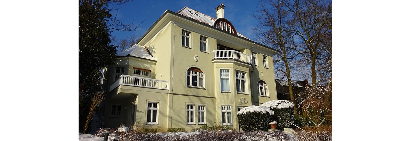 Garden view of the guest house in winter, with a blue sky and snow on the roof, trees and bushes
