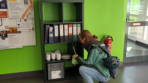Das Bild zeigt ein grünes Zimmer mit einem schwarzen Regal, auf dem Informationsmaterial und Ordner stehen. Eine Person hockt vor dem Regal und scheint etwas zu suchen oder zu betrachten, während im Vordergrund ein Schild "Gebraucht & Gut – Neues Tauschregal" zu sehen ist.