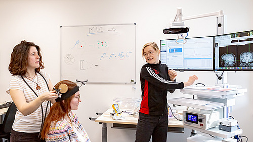 Drei Frauen arbeiten gemeinsam in einem Labor. Eine sitzt auf einem Stuhl, eine weitere erkl&auml;rt eine Computergrafik. 