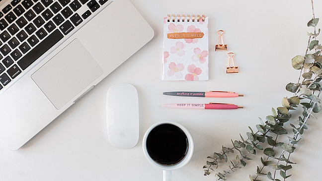 Ein Schreibtisch von oben gesehen. Links liegt ein aufgeklappter Laptop, daneben eine Computermaus.  In der Mitte liegen ein kleiner Notizblock mit Blumenmuster, zwei Stifte und zwei Metallklammern. Auf dem Notizblock steht &bdquo;Keep it simple&ldquo;.  Unten steht eine Tasse mit dunklem Getr&auml;nk. Rechts liegen Zweige mit runden Bl&auml;ttern. Der Tisch ist hell und aufger&auml;umt.