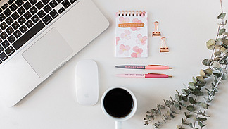 Ein Schreibtisch von oben gesehen. Links liegt ein aufgeklappter Laptop, daneben eine Computermaus.  In der Mitte liegen ein kleiner Notizblock mit Blumenmuster, zwei Stifte und zwei Metallklammern. Auf dem Notizblock steht &bdquo;Keep it simple&ldquo;.  Unten steht eine Tasse mit dunklem Getr&auml;nk. Rechts liegen Zweige mit runden Bl&auml;ttern. Der Tisch ist hell und aufger&auml;umt.