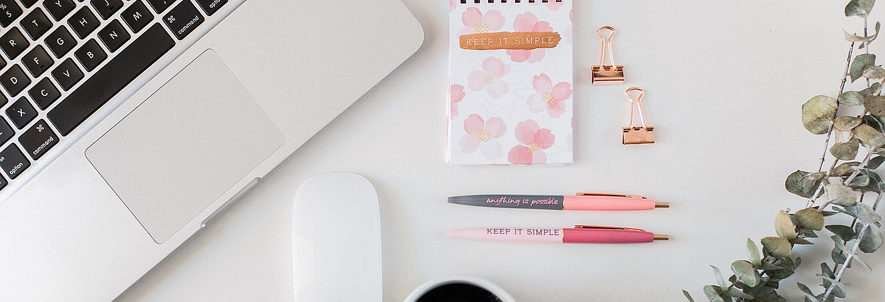 Ein Schreibtisch von oben gesehen. Links liegt ein aufgeklappter Laptop, daneben eine Computermaus.  In der Mitte liegen ein kleiner Notizblock mit Blumenmuster, zwei Stifte und zwei Metallklammern. Auf dem Notizblock steht &bdquo;Keep it simple&ldquo;.  Unten steht eine Tasse mit dunklem Getr&auml;nk. Rechts liegen Zweige mit runden Bl&auml;ttern. Der Tisch ist hell und aufger&auml;umt.