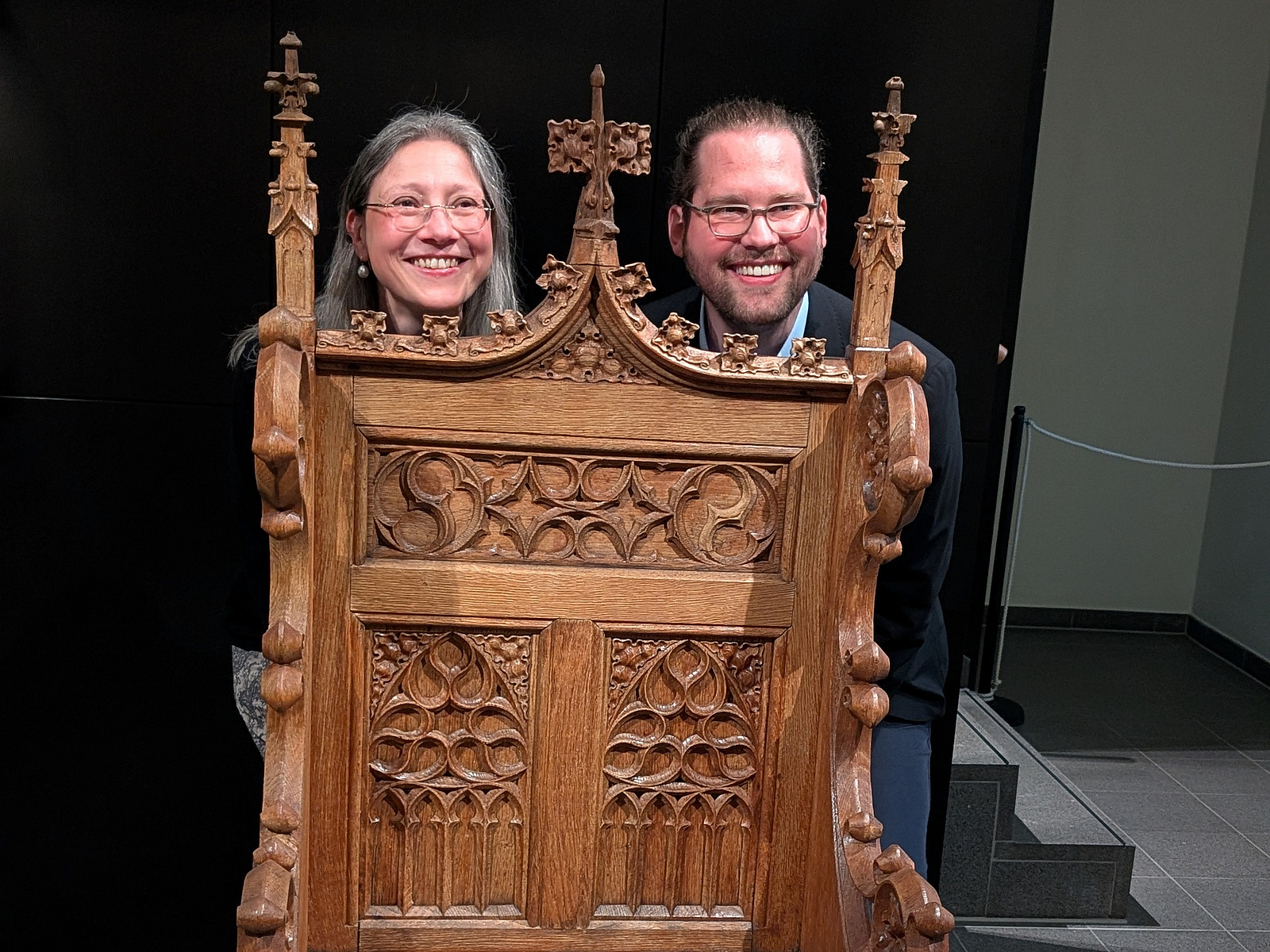 Zwei Personen schauen durch &Ouml;ffnungen in einem reich verzierten Holzobjekt. Das Objekt ist dunkelbraun, mit Schnitzereien und kleinen spitzen Aufs&auml;tzen verziert.  Die beiden Gesichter sind nebeneinander sichtbar und blicken in Richtung Kamera. Im Hintergrund ist eine dunkle Fl&auml;che zu sehen.