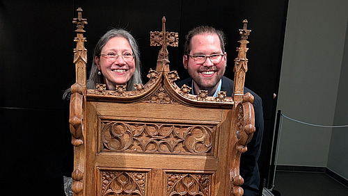 Zwei Personen schauen durch &Ouml;ffnungen in einem reich verzierten Holzobjekt. Das Objekt ist dunkelbraun, mit Schnitzereien und kleinen spitzen Aufs&auml;tzen verziert.  Die beiden Gesichter sind nebeneinander sichtbar und blicken in Richtung Kamera. Im Hintergrund ist eine dunkle Fl&auml;che zu sehen.