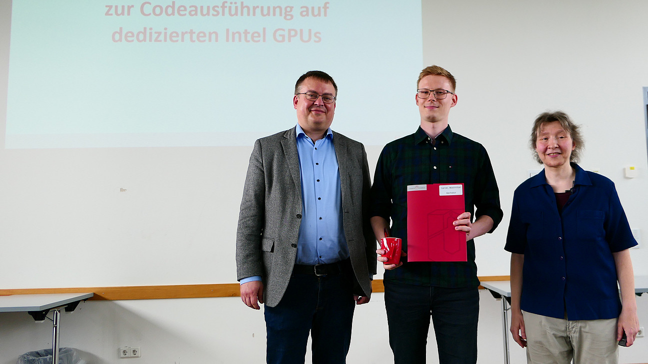 Ein Student mit Studiendekanin und Dekan stehen nebeneinander. Der Student hat eine Mappe in der Hand