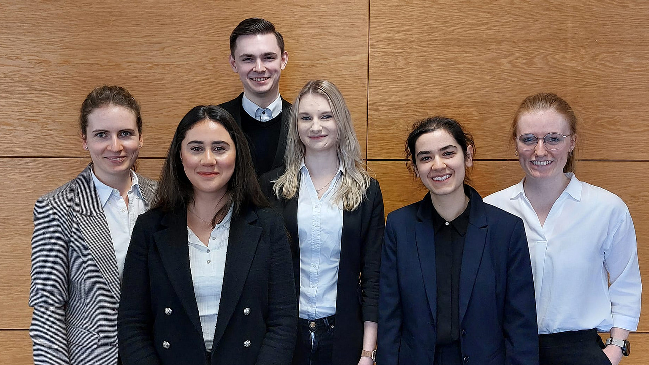 F&uuml;nf Frauen in zwei Reihen und ein gro&szlig;er Mann in der letzten Reihe vor einer braun vert&auml;felten Wand stehend 