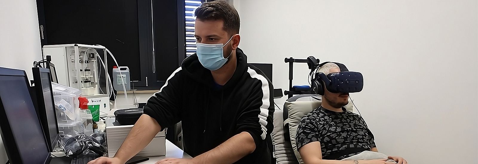 Das Bild zeigt eine Szene in einem Labor oder Testumfeld, in dem eine Person mit einem Virtual-Reality-Headset in einem gemütlichen Sessel sitzt. Die Person trägt Kopfhörer und ist entspannt, mit einem Kissen auf dem Schoß, was auf eine komfortable Testumgebung hinweist. Rechts neben dem sitzenden Probanden ist eine zweite Person zu sehen, die am Schreibtisch an einem Computer arbeitet und auf den Monitor schaut.