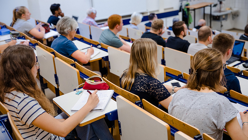 View from behind across rows of seats in a lecture hall to students, including elderly people.