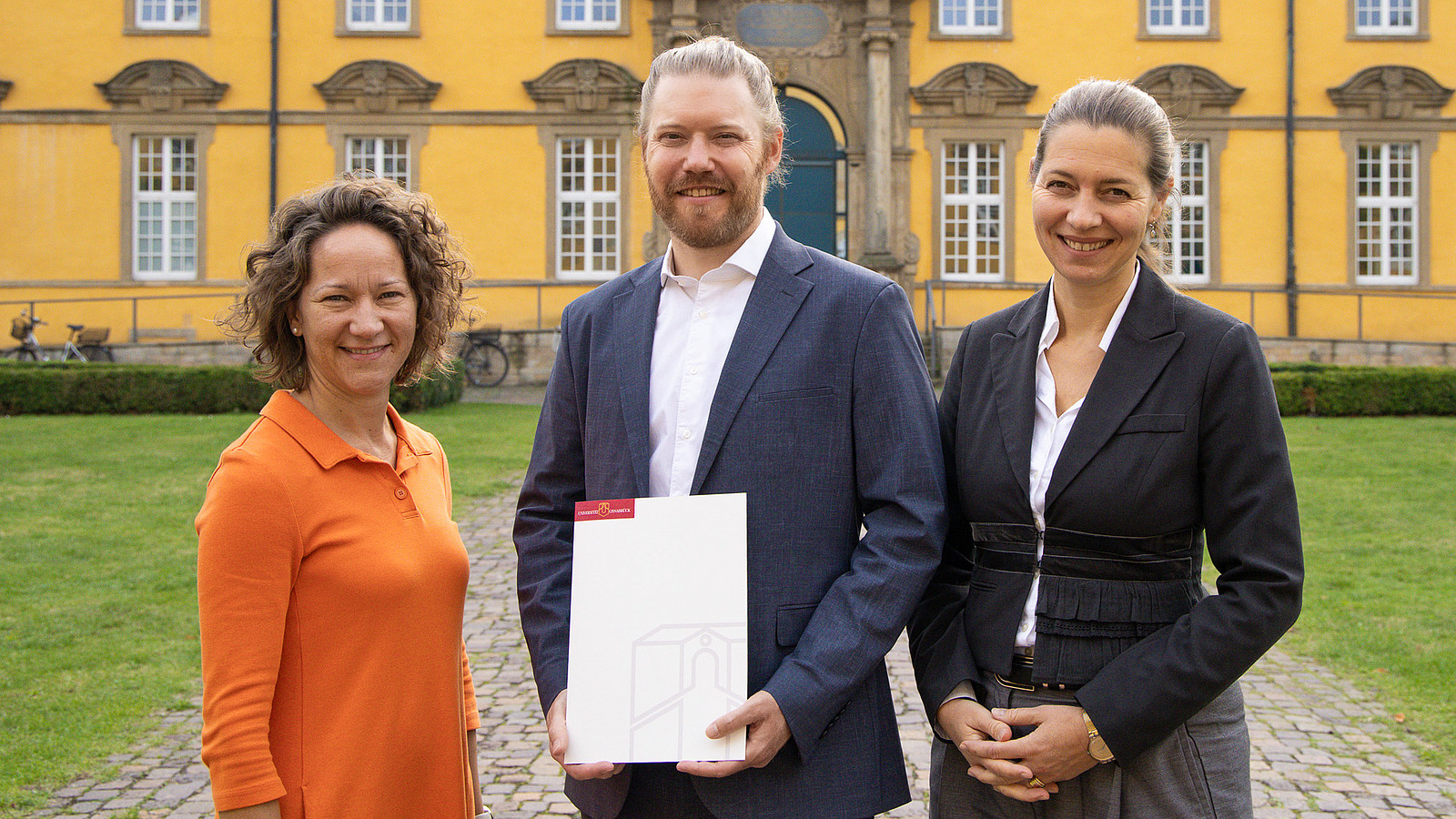 Drei lächelnde Personen stehen vor dem historischen Gebäude der Universität Osnabrück. 