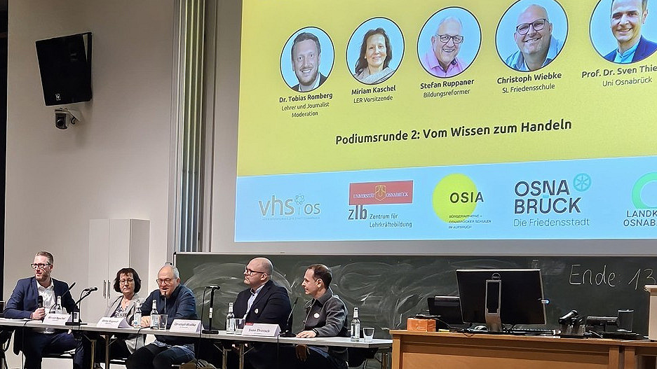 Men and women sit at a table on a podium and discuss. 