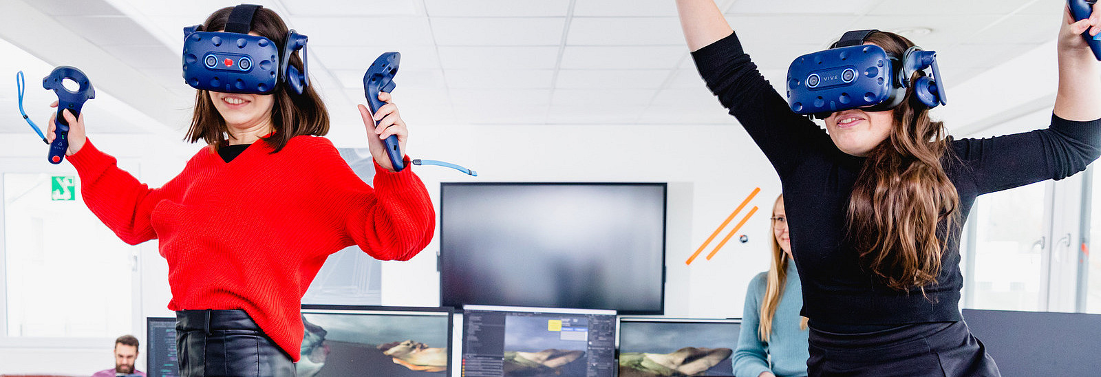 Two young women are jumping in the air in a modern office while wearing virtual reality headsets and holding controllers. They appear excited and focused. In the background, other people are working at computers, while a colleague watches them. The office is modern, with large screens, plants, and open workspaces.