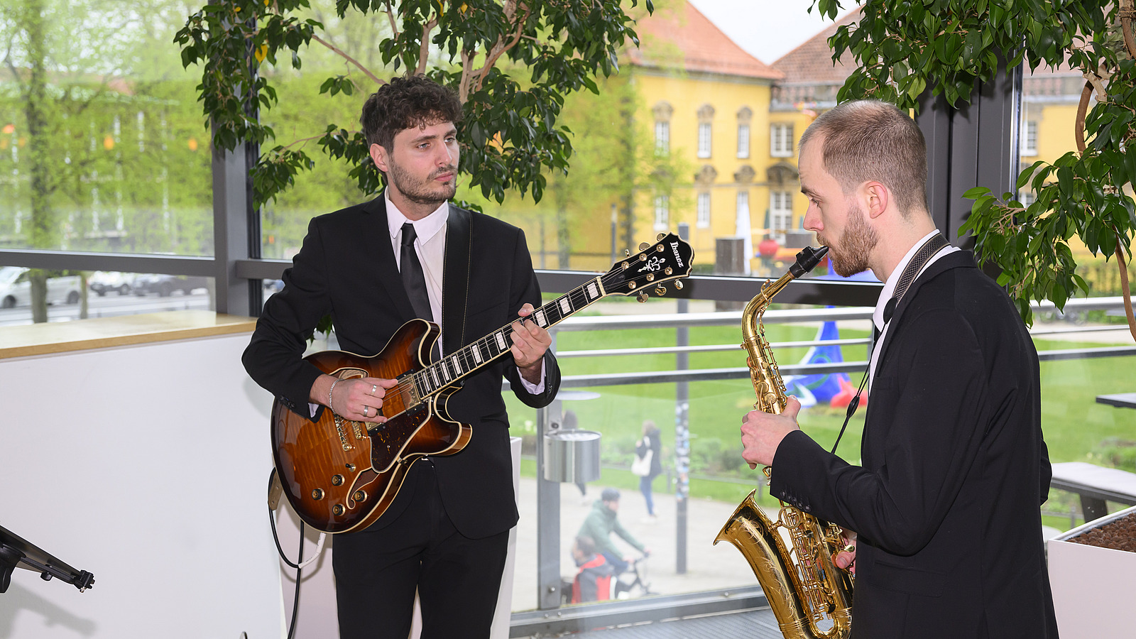 Two musicians with instruments in front of a large window, yellow castle building in the background